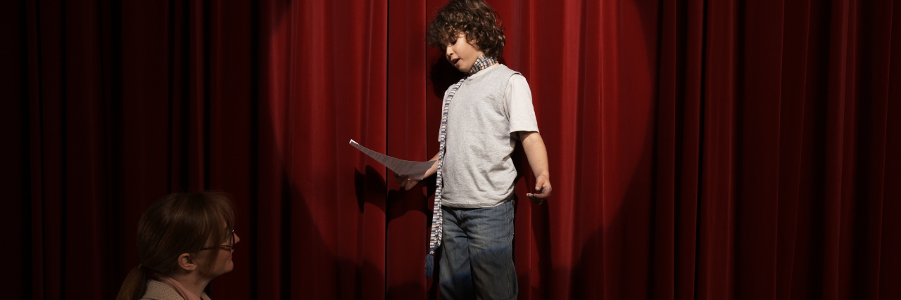 Boy acting on stage as teacher watches.