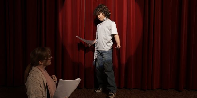 Boy acting on stage as teacher watches.