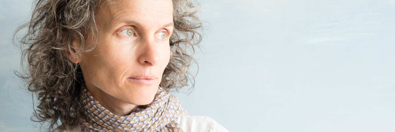 Invisible Illness: What It Feels Like to Live With Fibromyalgia woman wearing a white shirt and scarf standing against a light blue wall and looking away