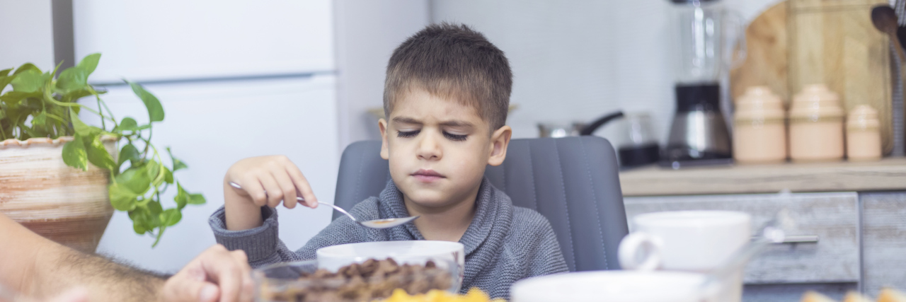 I'm an OT and I Put Chicken Nuggets On My Head Young boy at dinner table holding spoon and unhapy about the food in front of him as if he does not want to eat.