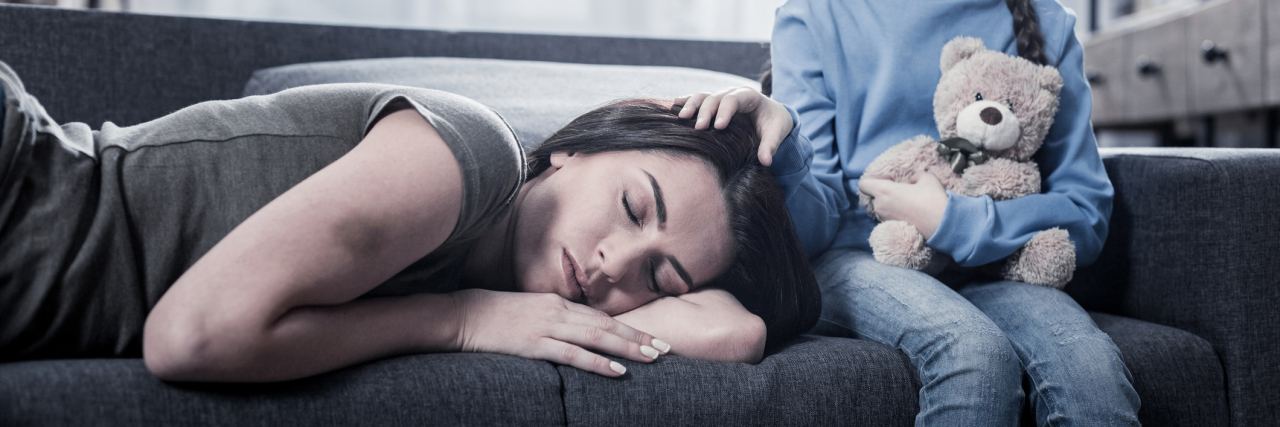 Why the Spoonie Parenting Struggle is Real Tired mom sleeping on the couch with daughter resting her hand on her mom's head.