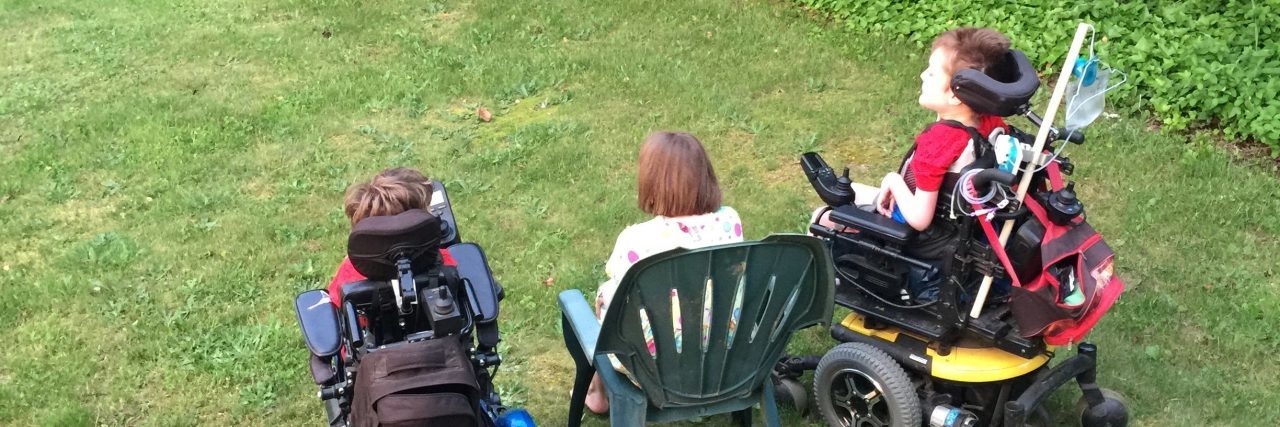 Back of two sons in wheelchairs, mom sitting on a lawn chair in the middle, they are all facing the street watching cars go by