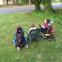 Back of two sons in wheelchairs, mom sitting on a lawn chair in the middle, they are all facing the street watching cars go by