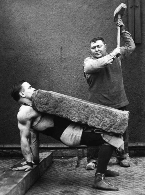 man with boulder on stomach and another man holding up hammer