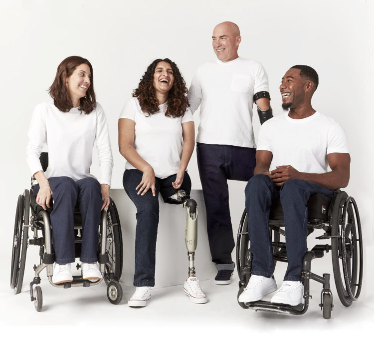 Four models wearing IZ Adaptive jeans. Two models are using wheelchairs. Two others with amputations are sitting and standing between them. All are wearing white t-shirts.