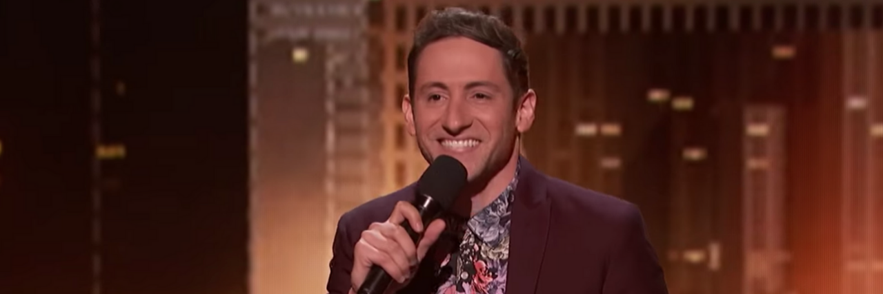Comedian With Tourette's Samuel Comroe May Win 'America's Got Talent' Samuel Comroe smiling while performing wearing a dark red blazer.