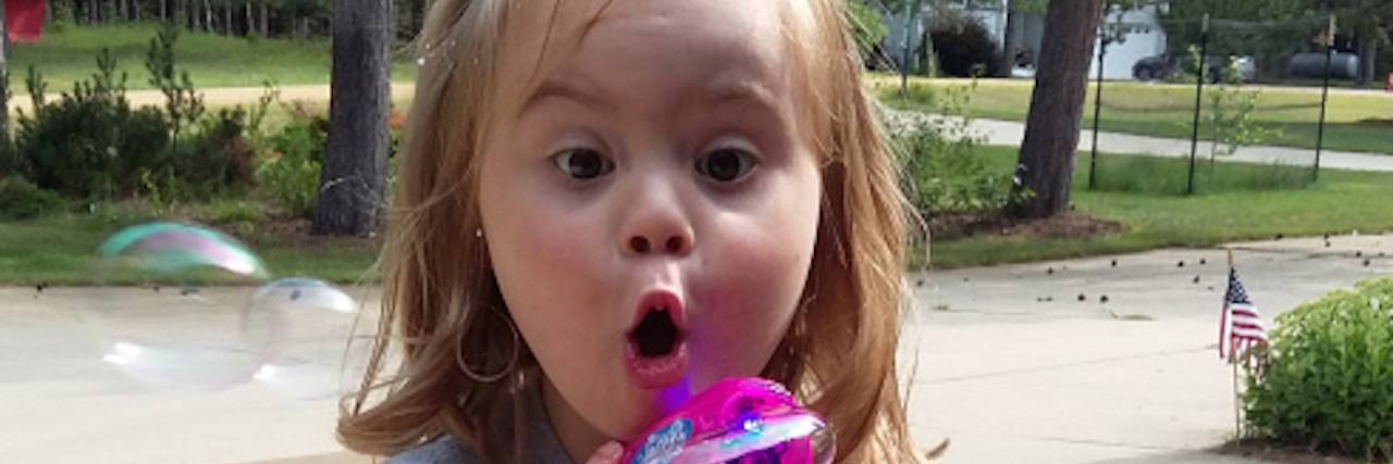 Little girl with Down syndrome holding a bubble maker and surprised expression on her face with big eyes and open mouth.