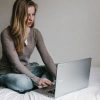 young woman sitting on bed typing on laptop