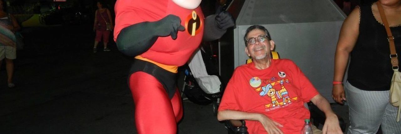 The Caregiver Self-Care Tip I Learned From Disney Movies Person in a red shirt and shorts smiling from a power wheelchair while posing with Mr. Incredible at night in a theme-park setting.