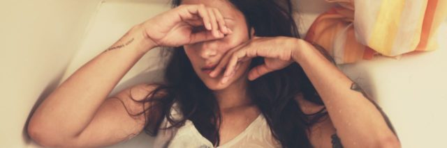 photo of woman with dark hair in bath looking tired or sad with hand partially covering eyes. She's wearing a white shirt