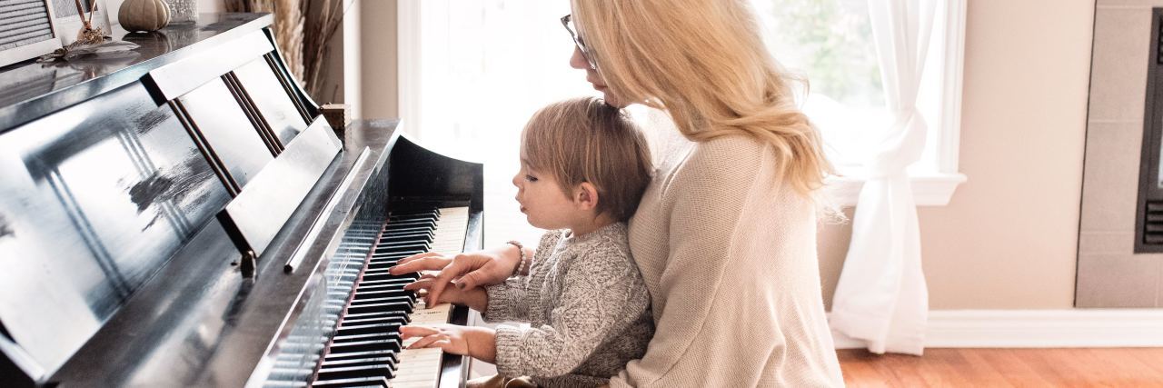 photo of young mother sitting at piano with young son in front of light-filled window while they play together