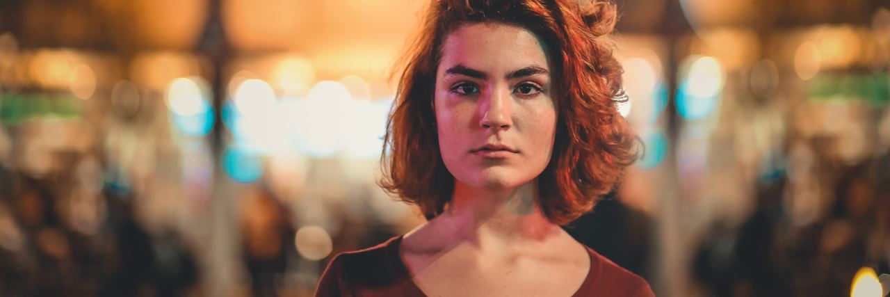 The Truth About Living With Chronic Illness woman with serious expression standing in front of blurred lights and outdoor activity