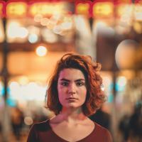 woman with serious expression standing in front of blurred lights and outdoor activity