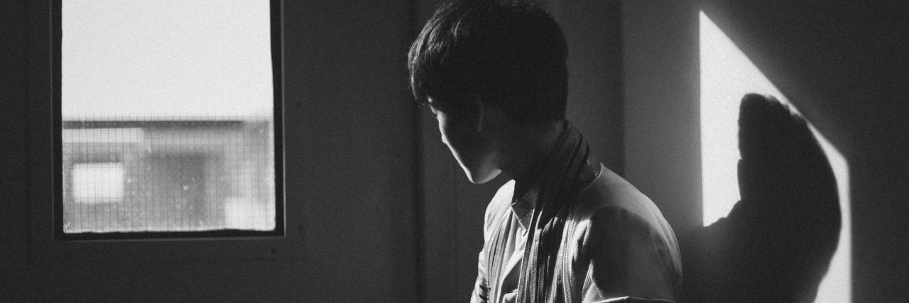 Can You Be Both a Survivor and a Victim With PTSD black and white photo of young man standing facing window with shadow behind him