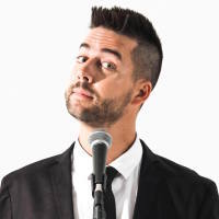 A photo of comedian John Crist standing in front of a white backdrop and microphone.