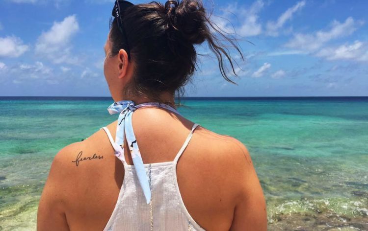A woman with the word fearless tattooed on her back. She's on the beach, looking out at the ocean, her dark hair in a bun.