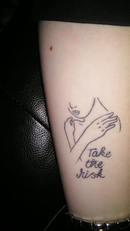 A tattoo of a woman with her arms around herself. Text underneath the figure reads: Take the risk