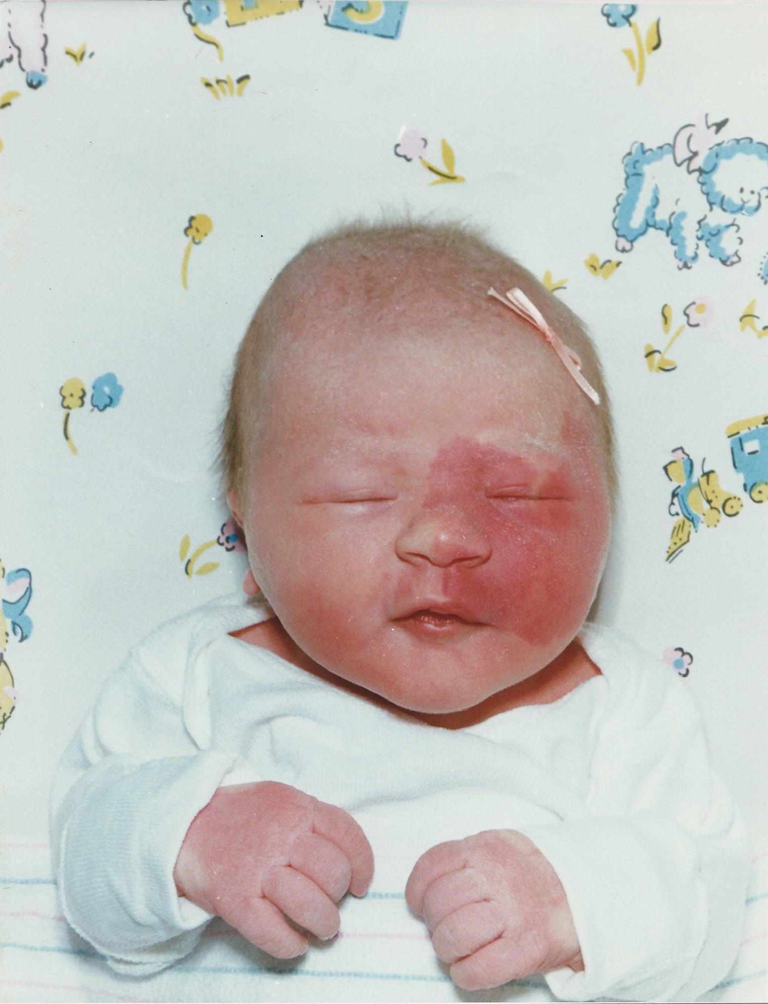 A newborn photo of Crystal, wearing a pink bow in her hear laying on a baby patterned blanket.