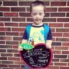 3-year-old with Down syndrme wearing a backpack on fist day of school holding a sign for his first day.