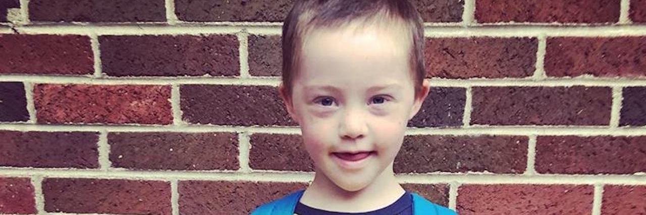 If My Child Had Been Born Without Down Syndrome 3-year-old with Down syndrme wearing a backpack on fist day of school holding a sign for his first day.