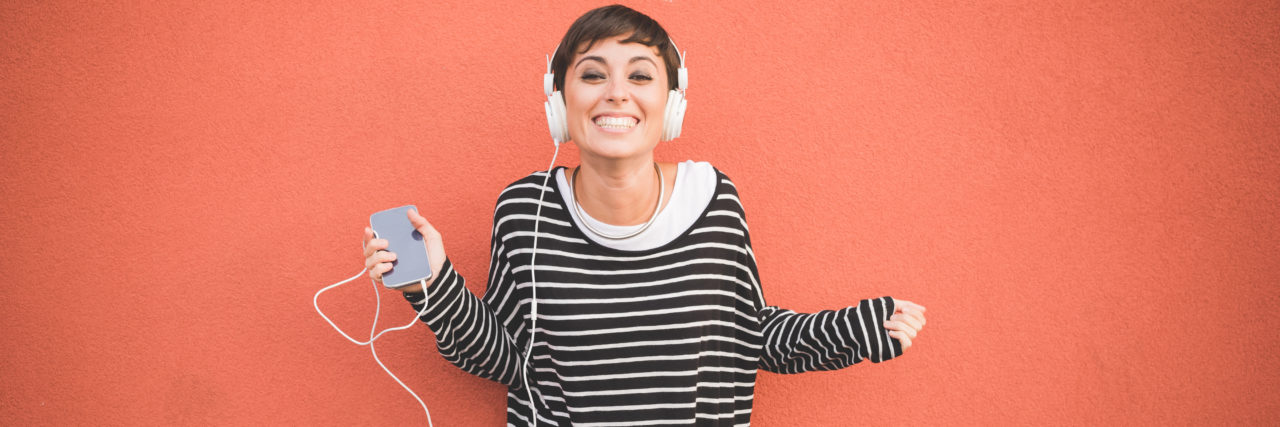 Tips for Coping and Thriving as an Adult With Autism and Anxiety Half length of young handsome caucasian brown straight hair woman listening music with headphones leaning against a wall, looking in camera smiling, holding smartphone - technology, music concept