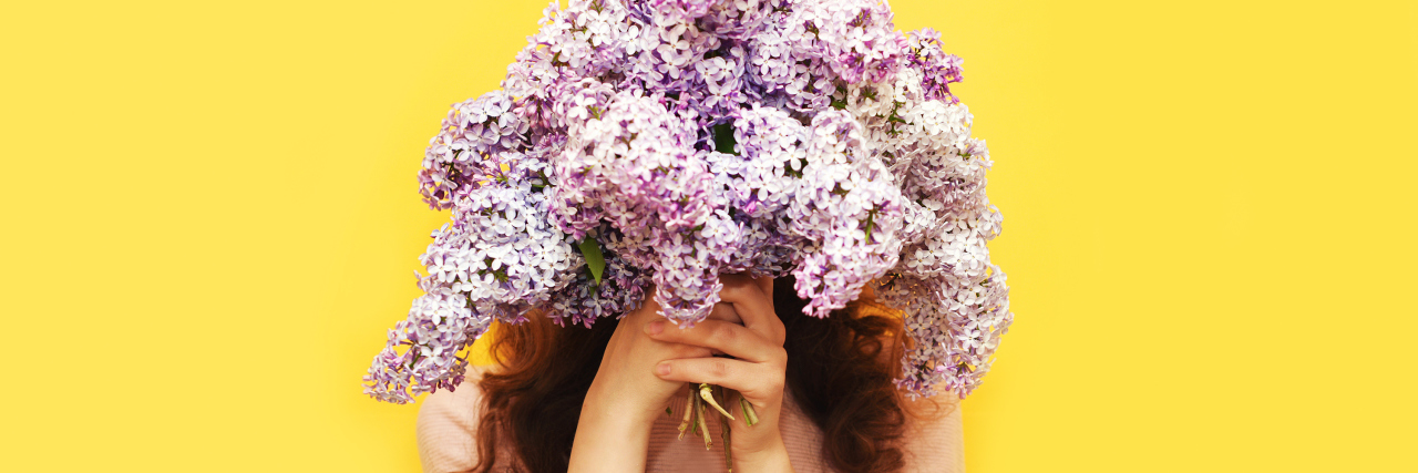 Woman hiding head in bouquet lilac flowers over yellow background