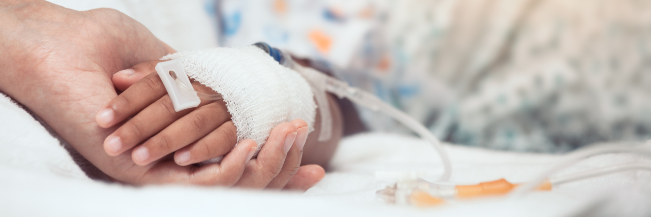 A parent holding the hand of a child who is receiving IV fluid and is in a hospital bed.
