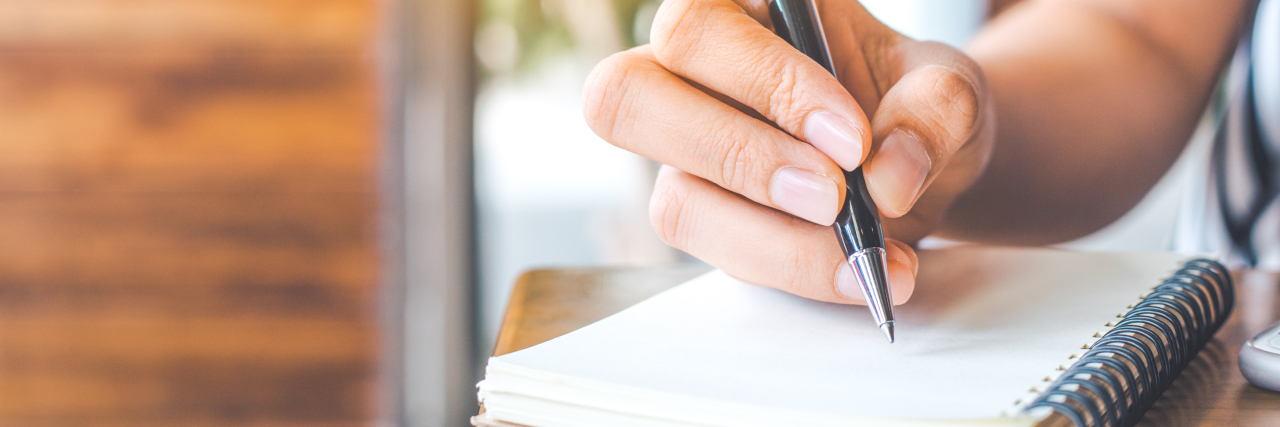 On the Days Life Stands Still With My Daughter's Rare Disease, I Write Myself This Letter woman's hand is writing on a blank notepad with a pen on a wooden desk.