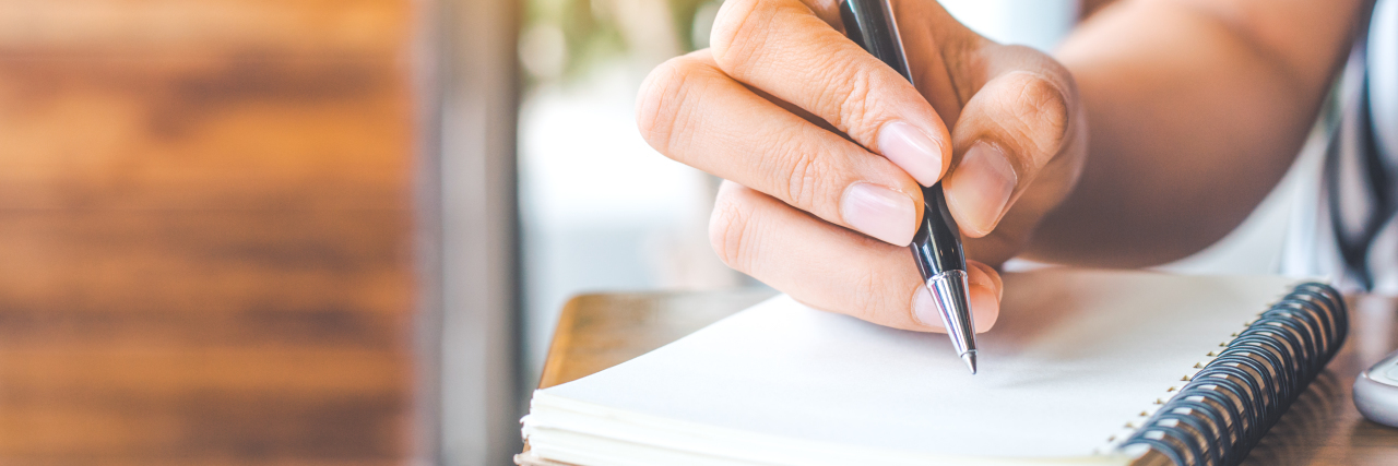 woman's hand is writing on a blank notepad with a pen on a wooden desk.