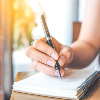 woman's hand is writing on a blank notepad with a pen on a wooden desk.