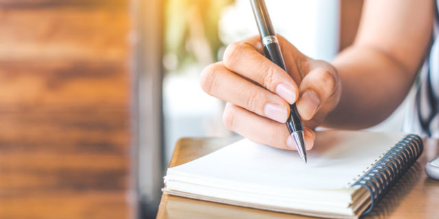 woman's hand is writing on a blank notepad with a pen on a wooden desk.