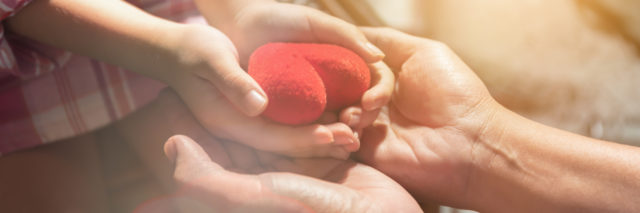 Adult hands and child's hands holding a red heart.