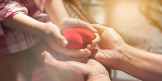 Adult hands and child's hands holding a red heart.