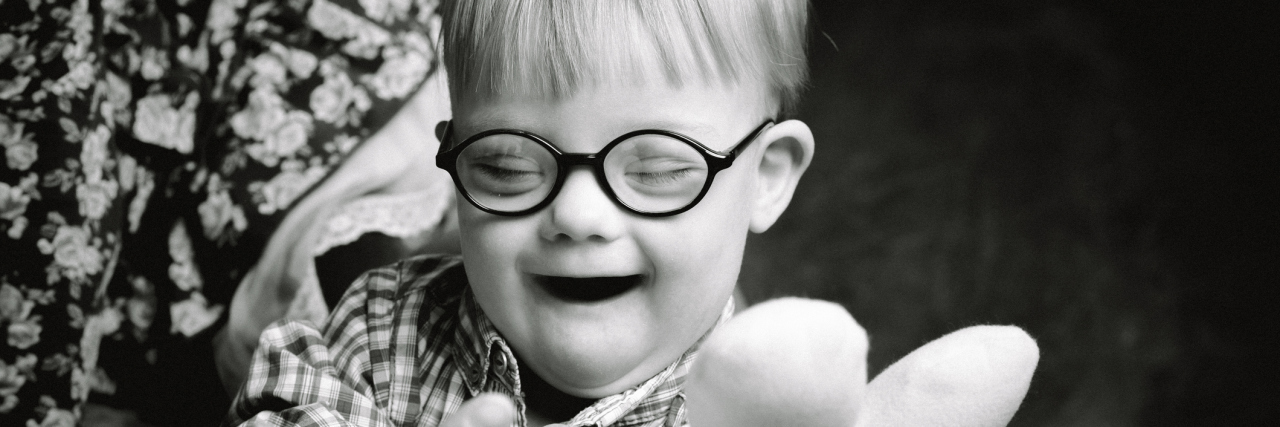 Kindness Is What My Son With Down Syndrome Needs Most From His Peers Black and white image of boy with Down syndrome playing with a stuffed toy, smiling, and sitting on a parent's lap.