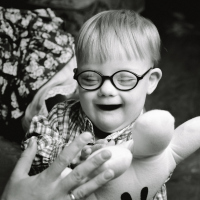 Black and white image of boy with Down syndrome playing with a stuffed toy, smiling, and sitting on a parent's lap.