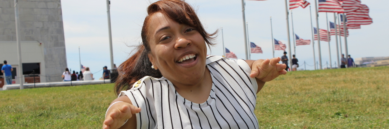 Living Well With Diastrophic Dysplasia Monique sitting in the grass with a row of American flags behind her.