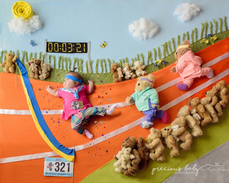 Baby with Down syndrome in a "race." She is about to cross the finish line with a bue and yellow ribbon, two teddy bears run behind her while other teddy bears watch on the sidelines. 