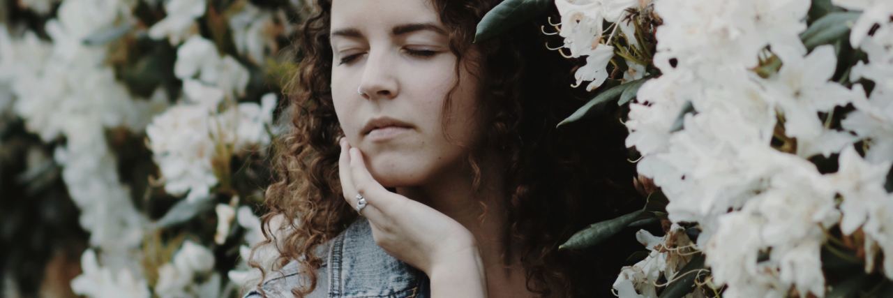 photo of young woman with brown curly hair standing by white flowers with eyes closed