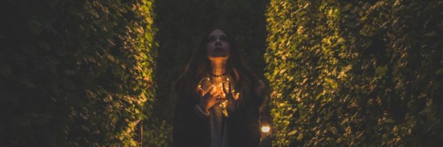 Woman standing between two hedges looking up while holding fairy lights