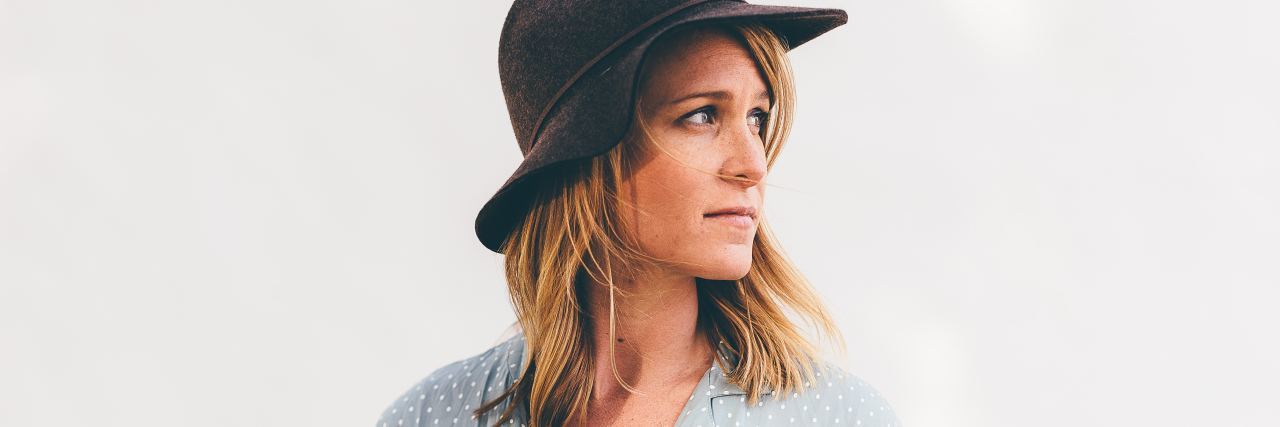 Deciding Whether to Reveal or Hide Your Chronic Illness woman with short blonde hair wearing a blue shirt and a brown hat is looking away over her shoulder and standing against a white wall