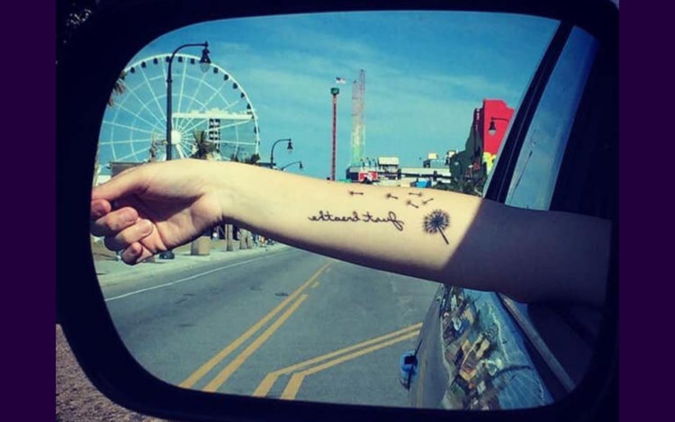 A woman holding her hand out the window, we can see the reflection in the side mirror. On her arm her tattoo reads: just breathe