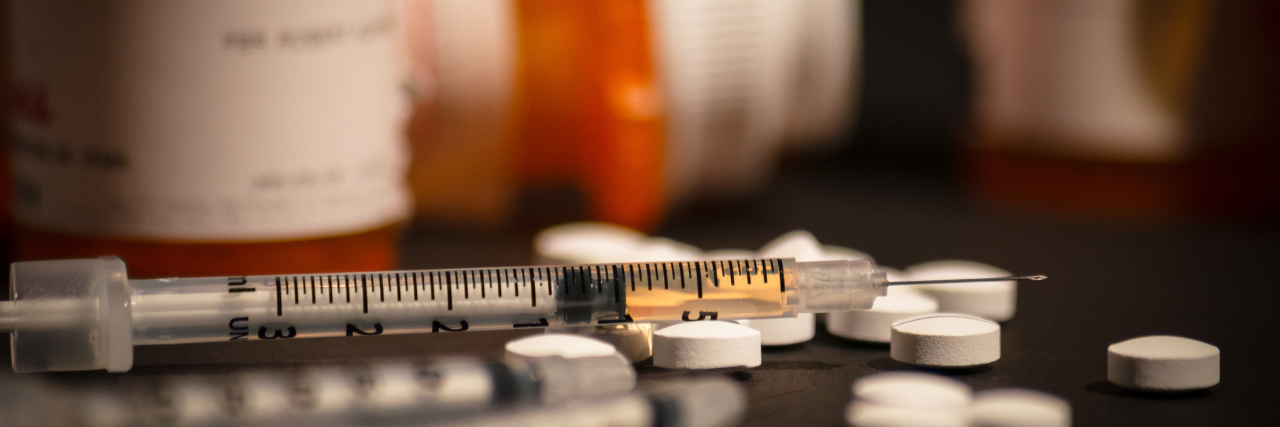 An injectable drug is loaded into a syringe while prescription medication is strewn about haphazardly.