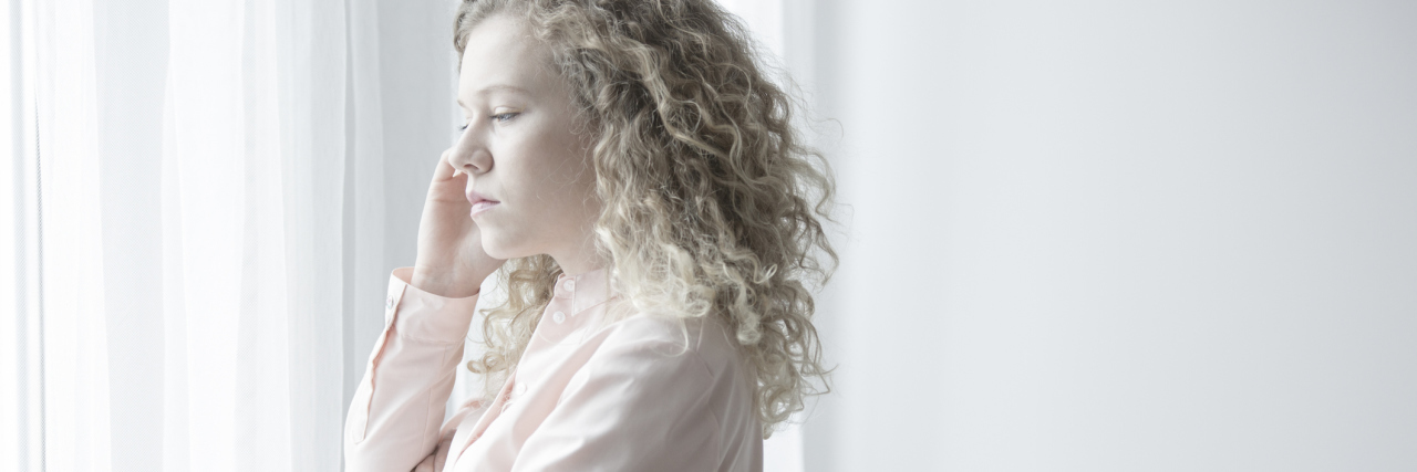 young blonde woman standing by window in room with white walls