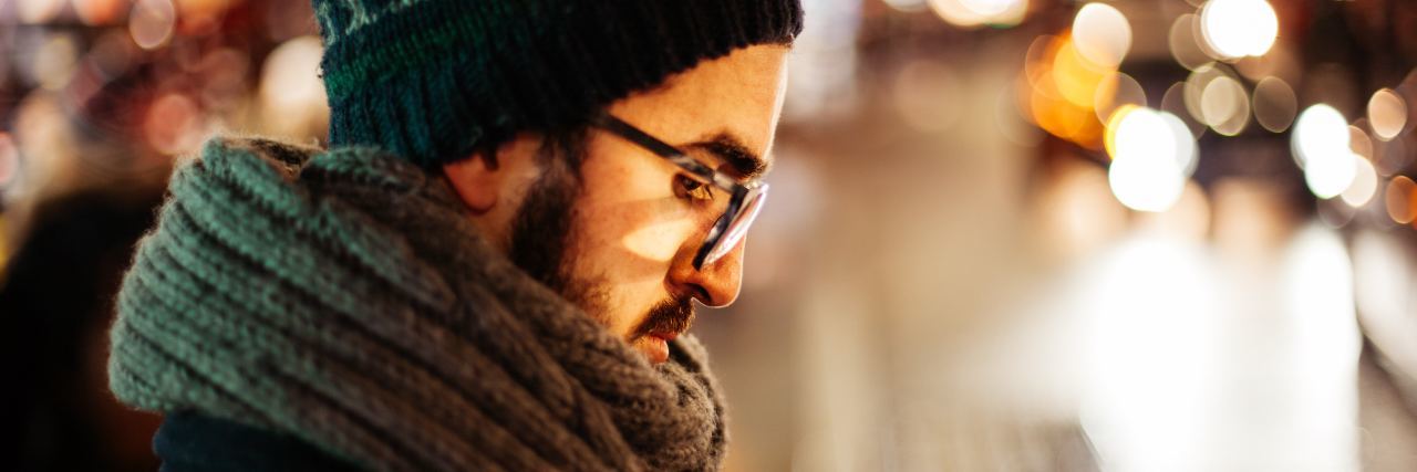 man with beard and glasses standing on street at night looking down and tired