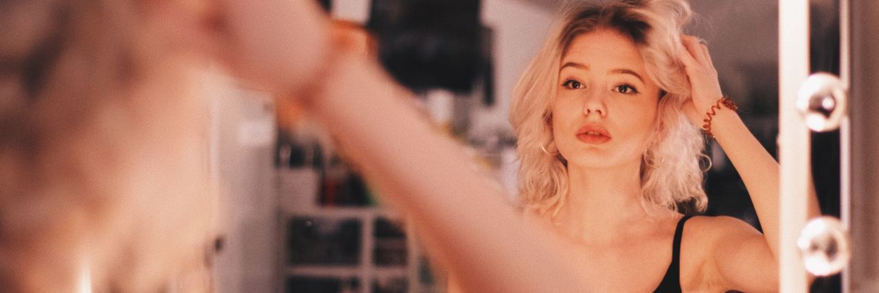 What You Need to Know About Trichotillomania and Related Disorders young blonde woman looking into mirror at reflection with hand in her hair