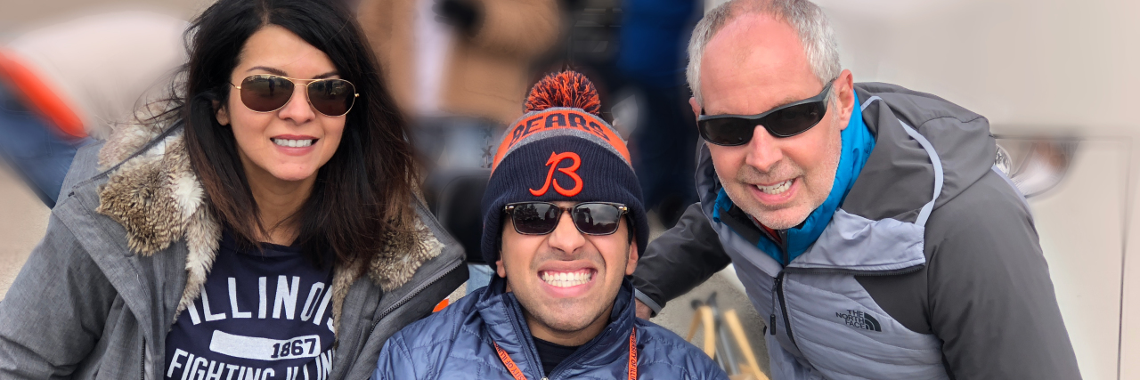 My Child With a Disability 'Belongs' Image of mother and father on each side of adult son who is sitting on his wheelchair. All three are smiling at the camera.