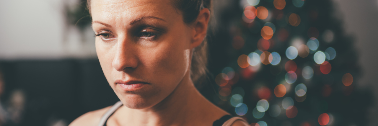 When Chronic Illness Affects Your Happiness Around the Holidays woman standing in front of a christmas tree looking sad