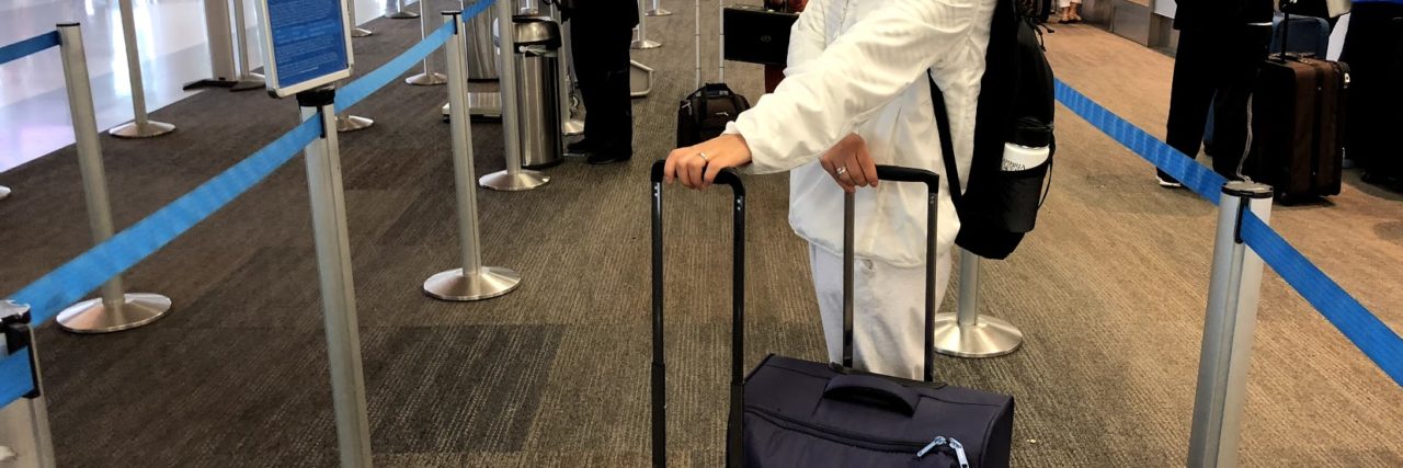 How to Survive the Holidays With Migraine the author standing in line at the airport with her suitcase. she's wearing a hat and sunglasses