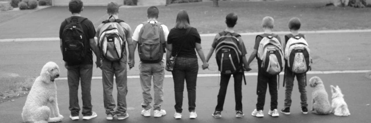 Why This Mom Adopted Five Kids With Down Syndrome Black and white image of Spring kids holding hands in a line across the driveway wearing backpacks, waiting for school bus