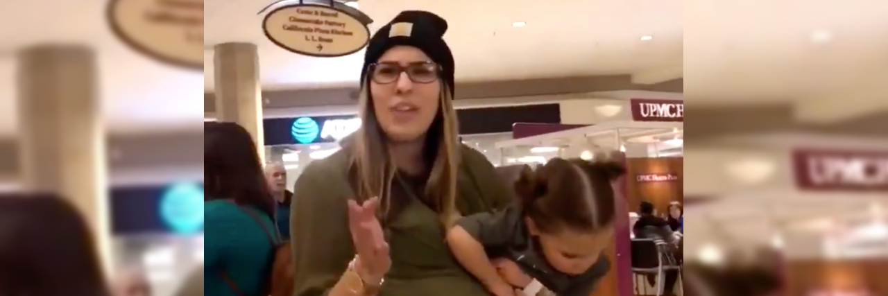 Mother holding her toddler daughter pointing at person filming. She's wearing a black beanie and a green blouse.